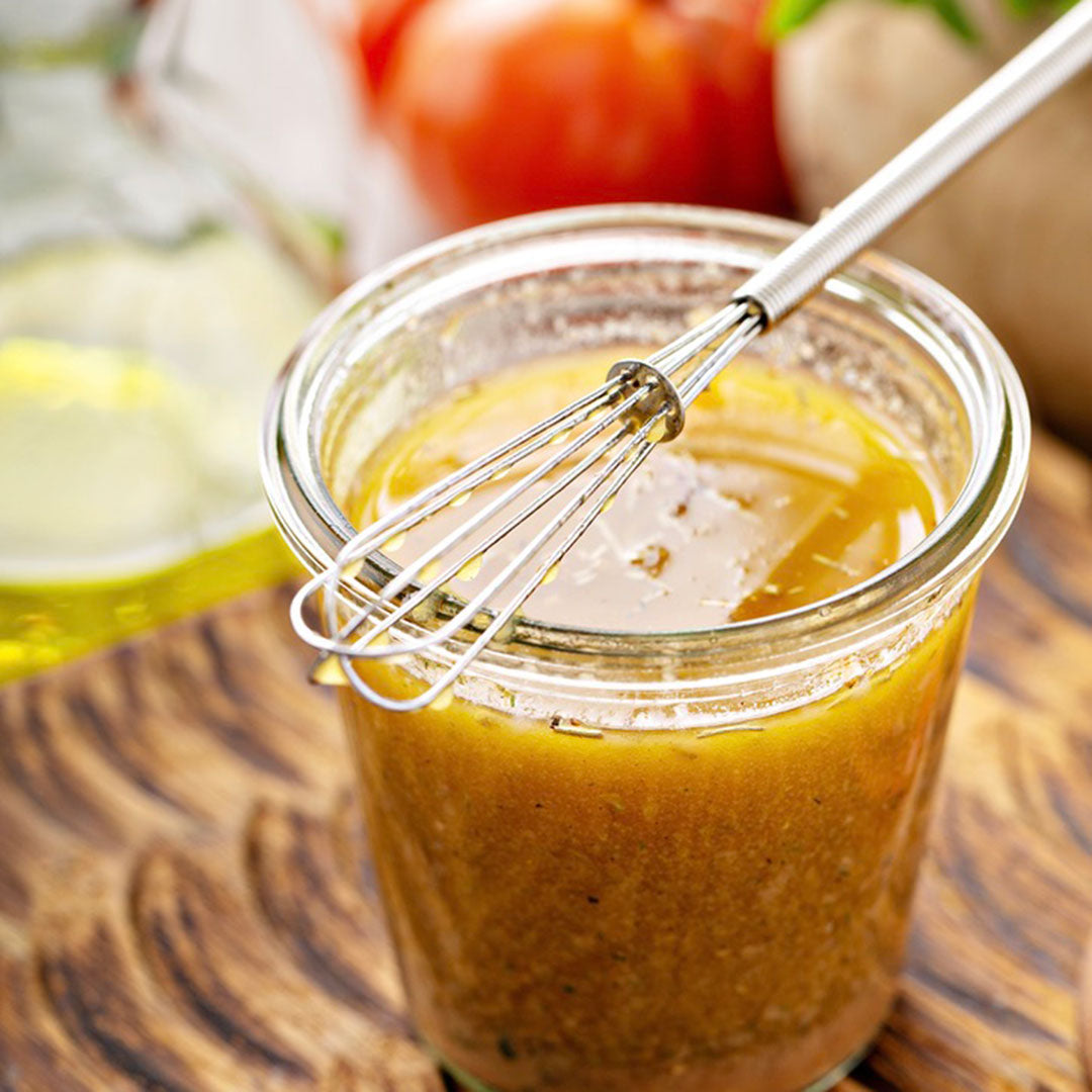 Glass jar with yellow dressing and a whisk, surrounded by tomatoes and a lemon on a wooden surface.