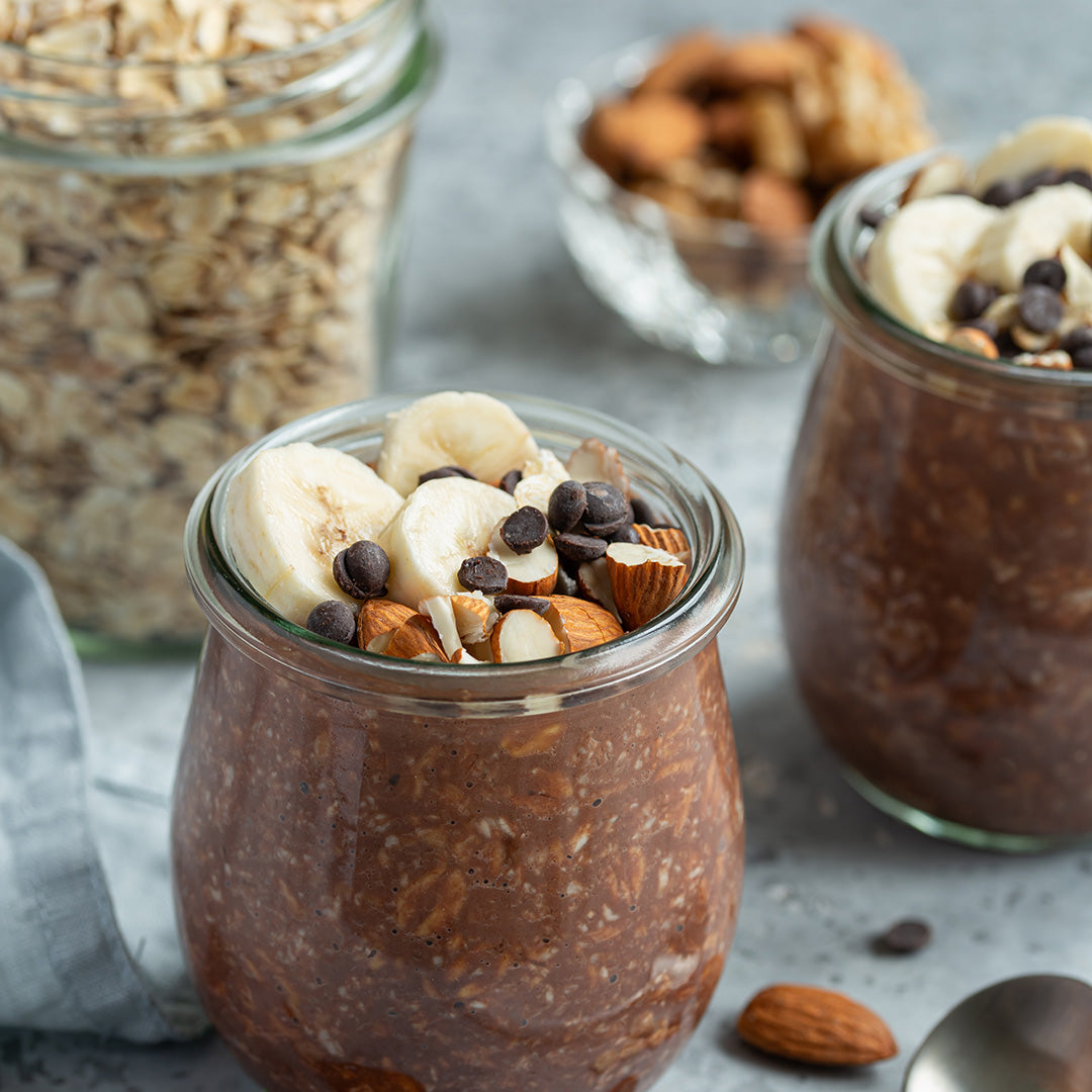Glass jars filled with chocolate pudding topped with bananas, nuts, and chocolate chips on a gray surface.