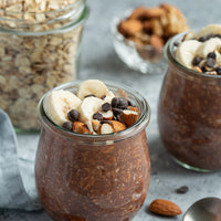 Glass jars filled with chocolate pudding topped with bananas, nuts, and chocolate chips on a gray surface.