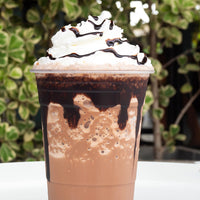 Chocolate drink with whipped cream and chocolate drizzle in a clear cup against a blurred green background
